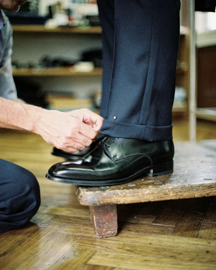 Sarto in ginocchio che appunta uno spillo sull'orlo di un pantalone blu dove tocca una scarpa nera, col cliente in piedi su una pedana di legno
