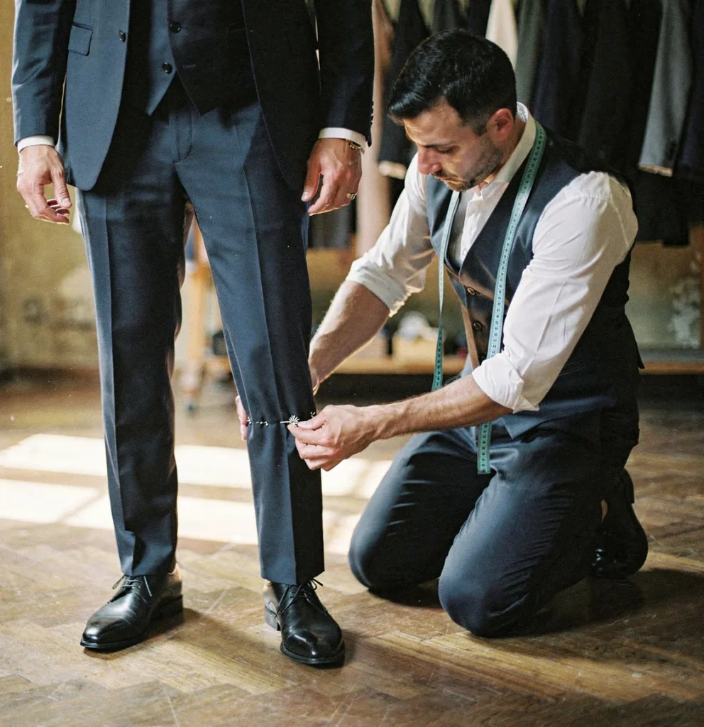 Sarto in ginocchio che appunta con gli spilli i pantaloni di un abito blu a tre pezzi indossato da un uomo, su un pavimento a spina di pesce